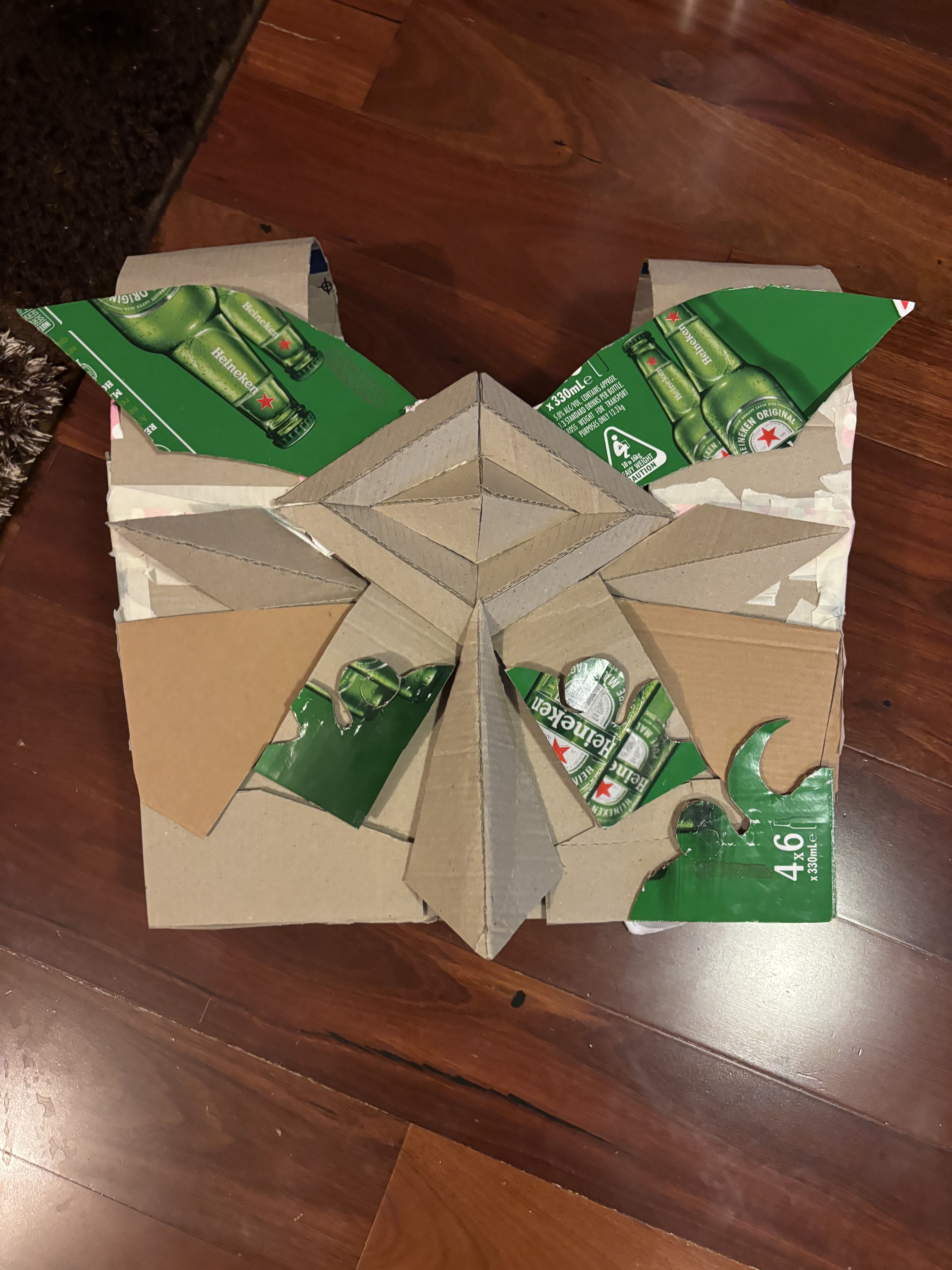 a handmade cardboard creation, using cardboard and heineken beer boxes, is displayed on a wooden floor. it appears to be a diy project or craft, potentially an artwork made from recycled materials.