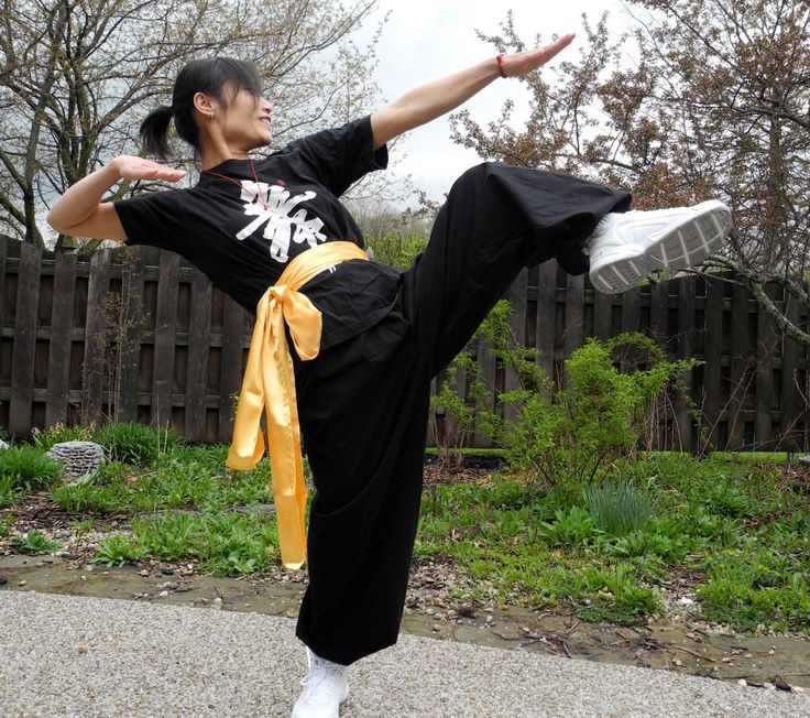 a woman in a black martial arts outfit and a yellow belt is executing a high kick in a garden setting. her right leg is extended and she's facing the camera. she's kicking toward the camera with a determined expression on her face. there are trees and grass in the background.