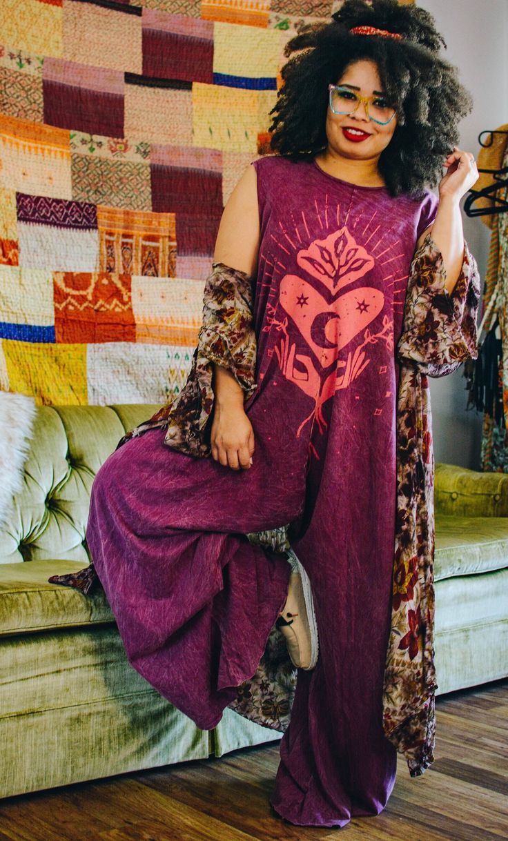 a woman with curly hair and wearing glasses is posing in a purple dress in front of a colorful quilt. she has one foot on a green couch. she is wearing a floral print scarf and jewelry. she is smiling and looking at the camera. she is also wearing casual sneakers.