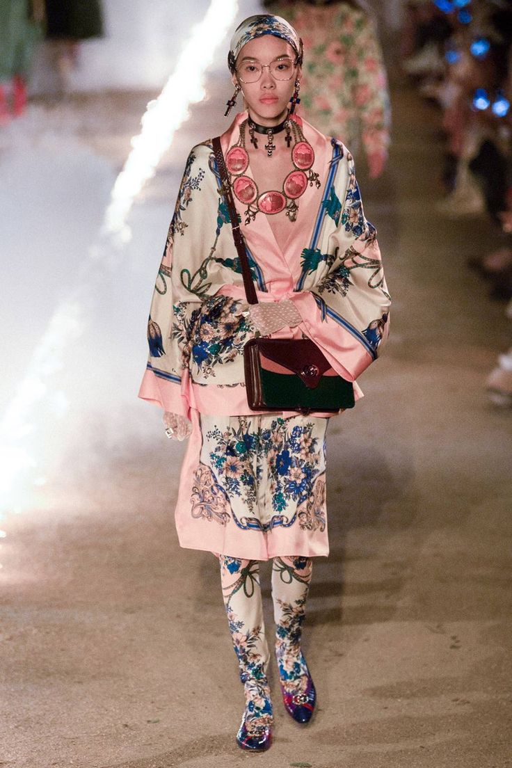 a model walks the runway at a gucci fashion show. she is wearing a pink and beige kimonostyle jacket and skirt with a floral print, paired with patterned tights and highheeled boots.  she carries a darkcolored handbag and is accessorized with glasses, a headscarf, and a statement necklace. the setting appears to be outdoors, with some atmospheric effects present. the overall aesthetic is one of high fashion and luxury.