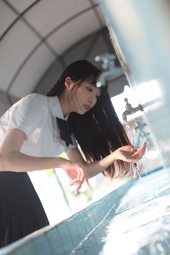 a young woman with long black hair wearing a white shirt and black skirt school uniform is washing her hands at a water fountain. the image is taken from a low angle and the background is blurred. the color palette is dominated by white, black, and blue.