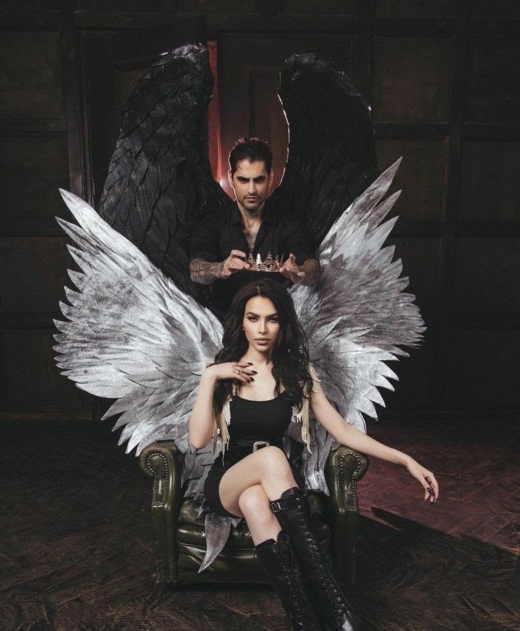 a woman with long black hair sits on a throne with white angel wings behind her. a man with black angel wings stands behind her and holds a crown above her head. the woman is dressed in a black dress and leather boots. she has a dark gothic look about her. the photo is set in a dark and moody setting.