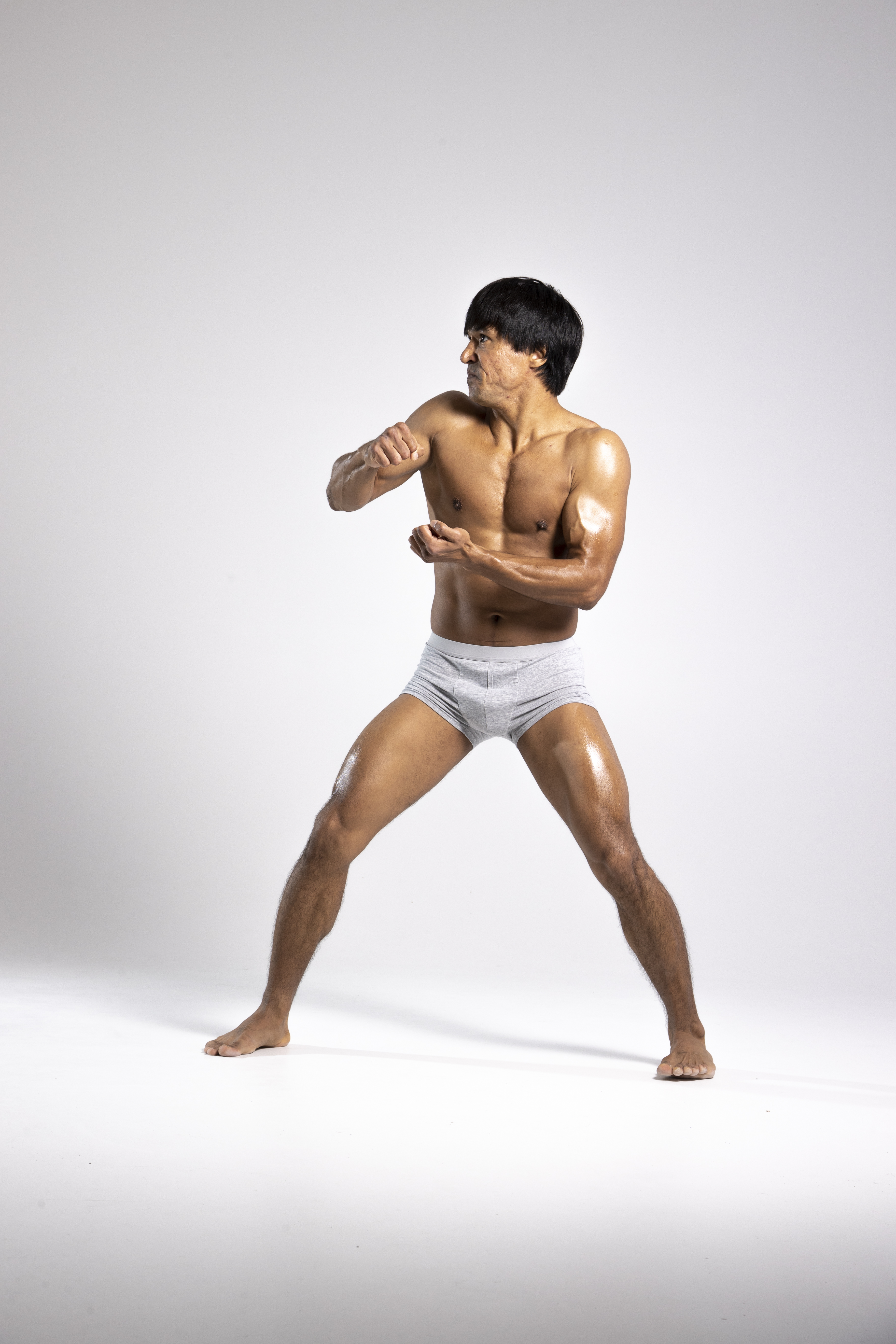 a shirtless man wearing gray shorts is in a fighting stance. he appears to be asian, has a muscular build, and his body has a sheen. he is facing to the side with his fists up. the background is a neutral gray.
