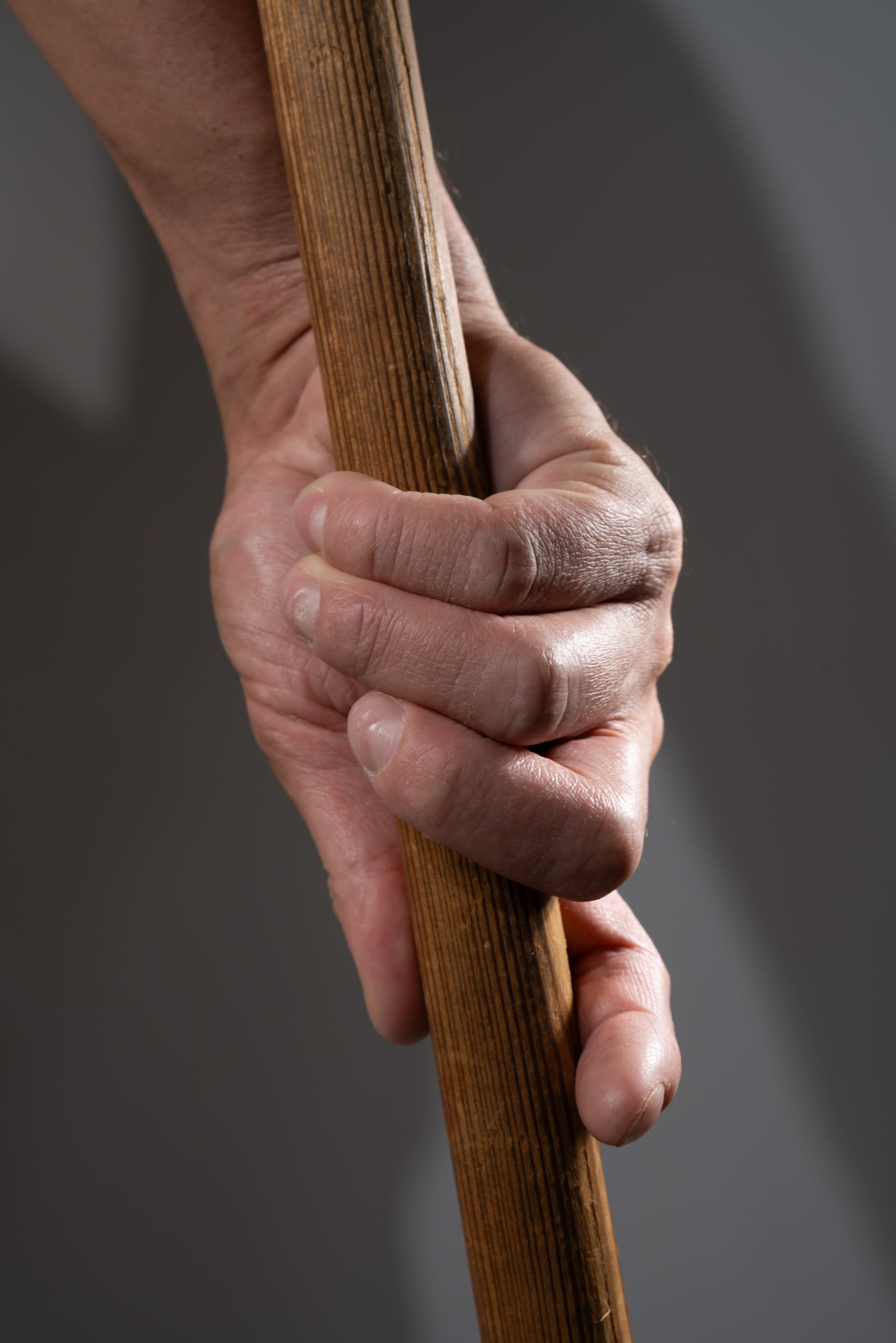 a closeup image of a man's hand tightly gripping a wooden stick. the texture of both the wood and the skin are highlighted by the studio lighting.