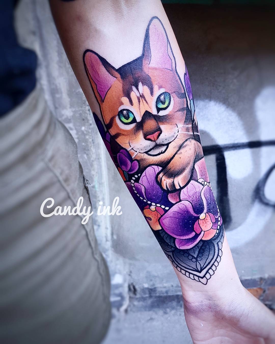 a person's left forearm with a colorful tattoo of a playful bengal kitten sitting in front of purple orchids. the tattoo has neotraditional elements with bold lines and bright colors. the background is blurred, and the focus is on the tattoo. the image is brightly lit.