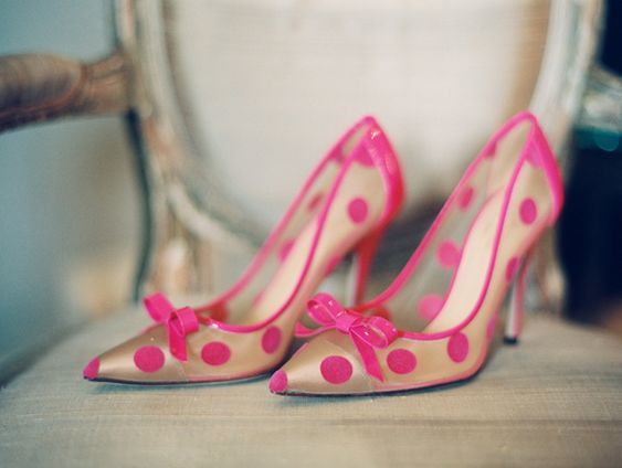 a pair of pink polka dot high heels with bows on the front. the shoes are clear and transparent. they are sitting on a light colored surface, possibly a chair.