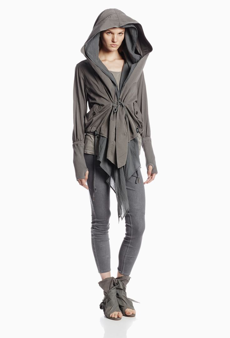 a woman is wearing a grey jacket with a hood and matching grey pants. she is posing in a studio with a white background.  she is wearing matching shoes that look like boots.  the woman's hood is up and the jacket is unbuttoned.  she is wearing a light colored shirt underneath the jacket.