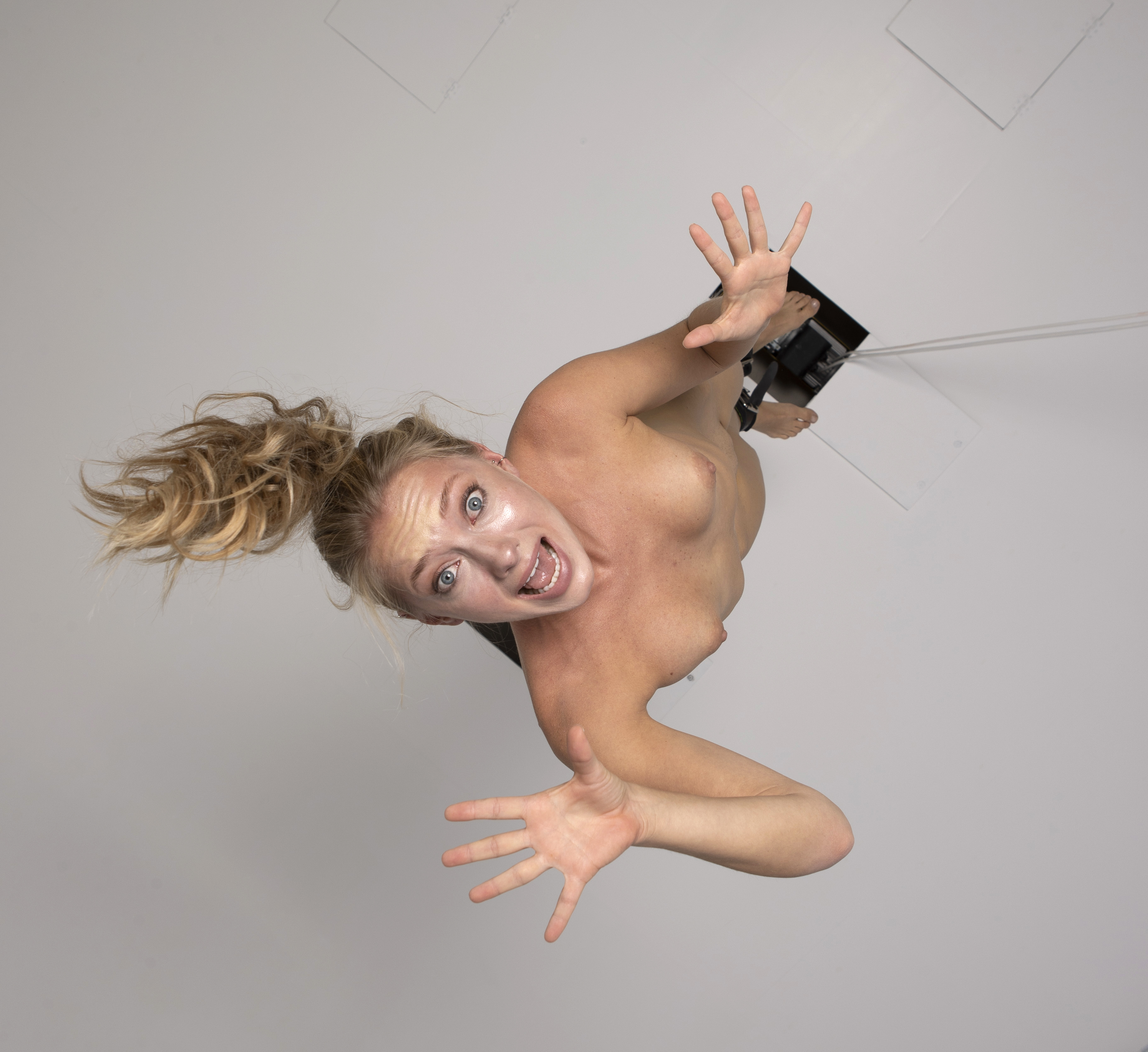 a nude woman with blonde hair in a ponytail is suspended from a harness attached to wires and ceiling tiles. she is looking directly at the camera with an open mouth and a surprised expression, her arms raised. the shot is taken from an overhead perspective in a studio setting.