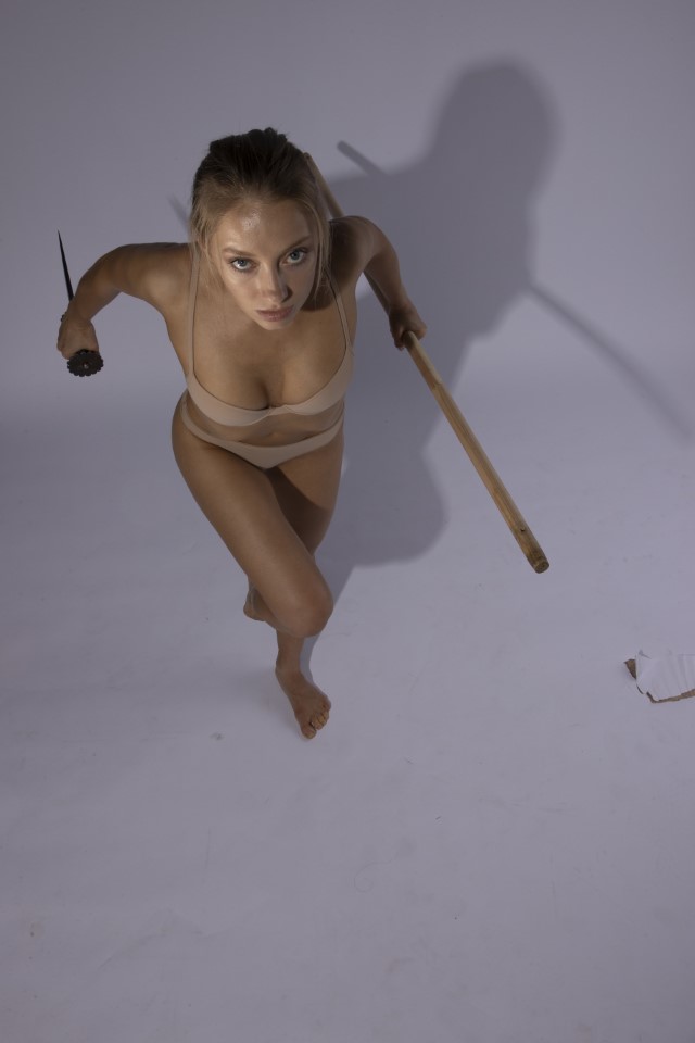 a full body shot of a young, fairskinned woman with blonde hair pulled back in a ponytail, running barefoot towards the camera. she is in a studio with a white background, holding a sword in her right hand and a knife in her left. she is looking up at the camera with an aggressive expression. the studio lighting creates a distinct shadow behind her.