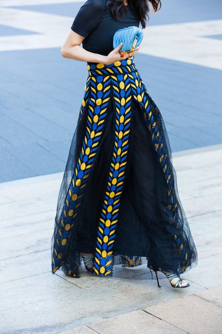 a woman wearing a black top and a long, flowing black skirt with intricate blue and yellow patterns is standing on a paved surface. she is holding a small blue clutch. the woman is wearing highheeled sandals.