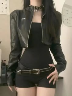 a young woman is wearing a black cropped leather jacket with a silver zipper, black shorts and a belt. she is posing in a studio setting.