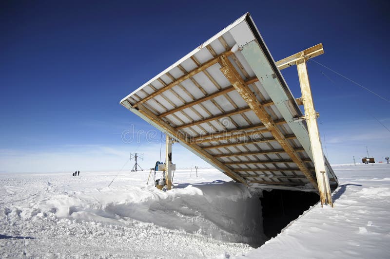 a metal door is open in a snowcovered landscape in antarctica.  the door is partially open, revealing a dark opening inside. the sky is bright blue and the snow is sparkling white. the door is a part of the research station.  a couple of people are walking away from the door,  in the distance.