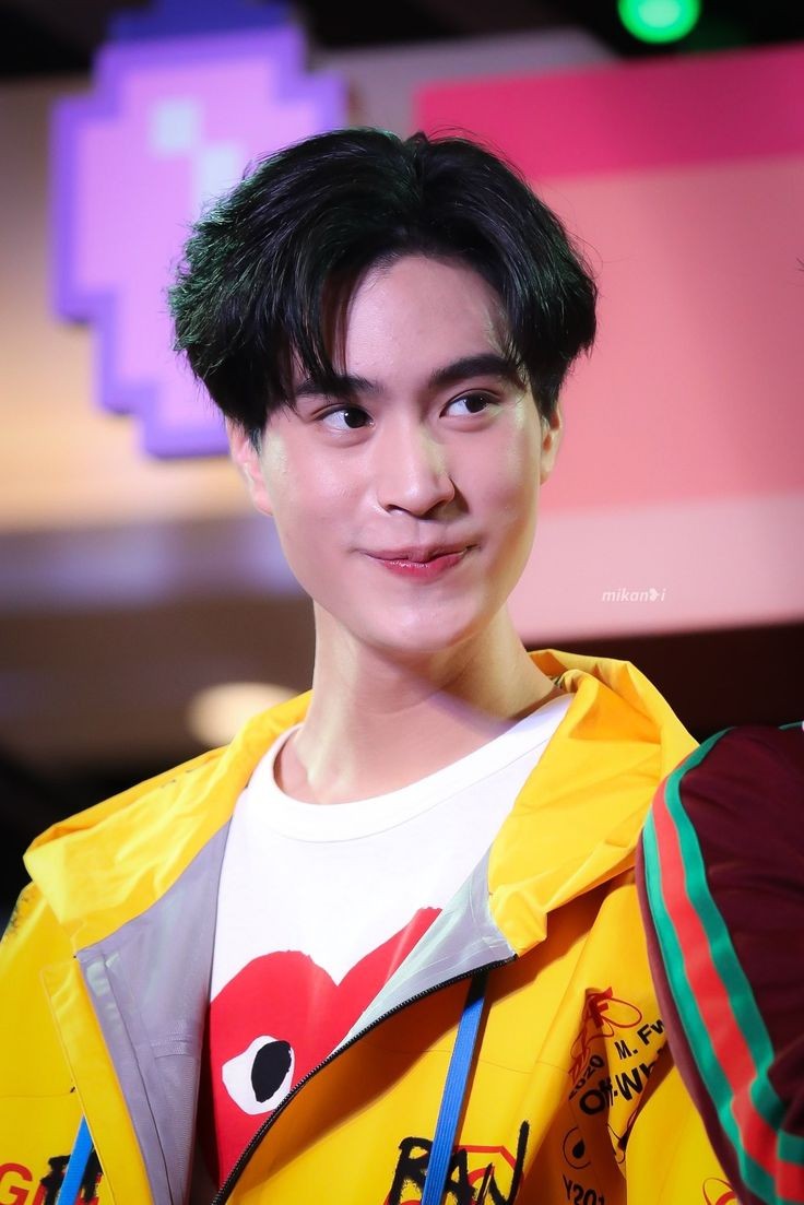 a closeup photo of a young man smiling. he has short, dark hair and is wearing a yellow jacket over a white shirt. the background is blurry and features shades of pink and purple.