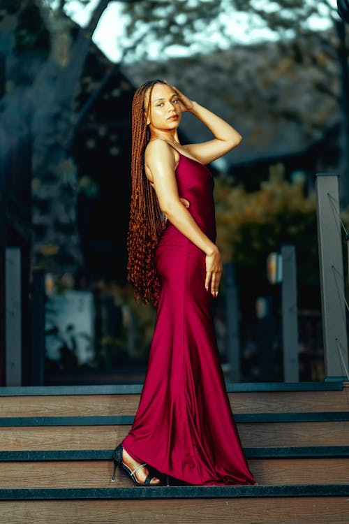 a stunning darkskinned woman with long brown braids is elegantly posed on a wooden staircase. she's wearing a flowing, crimsoncolored maxi dress that accentuates her figure.  the backdrop is a blurred, outdoor scene suggesting a luxurious or resortlike setting. the image is welllit, likely with natural light, giving it a warm and sophisticated feel.  the woman's confident pose and the overall aesthetic project a sense of elegance and style.