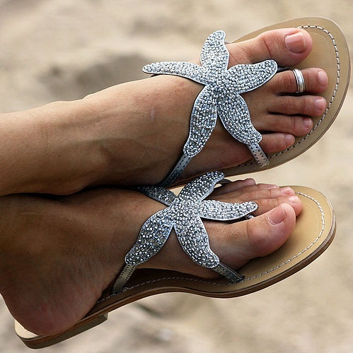 a closeup shot shows a pair of feet wearing flipflops adorned with silver starfish. the flipflops have a beige sole. a silver toe ring is visible on one of the toes. the background appears to be a sandy surface.