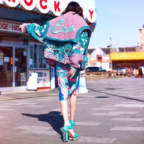 the image shows a fashion model from behind, wearing a stylish, oversized pastel pink and turquoise cardigan embroidered with sequins and the word "eiser." the cardigan is paired with a vibrant, patterned midi skirt in similar color scheme. the model is walking down a sunny street near a seaside boardwalk or amusement park. she is wearing bright turquoise platform heels and possibly sunglasses (though they are not clearly visible).  the overall style is casual yet highly fashionable and vibrant, capturing a summery and playful mood.  the background subtly suggests a beachside or amusement park environment.