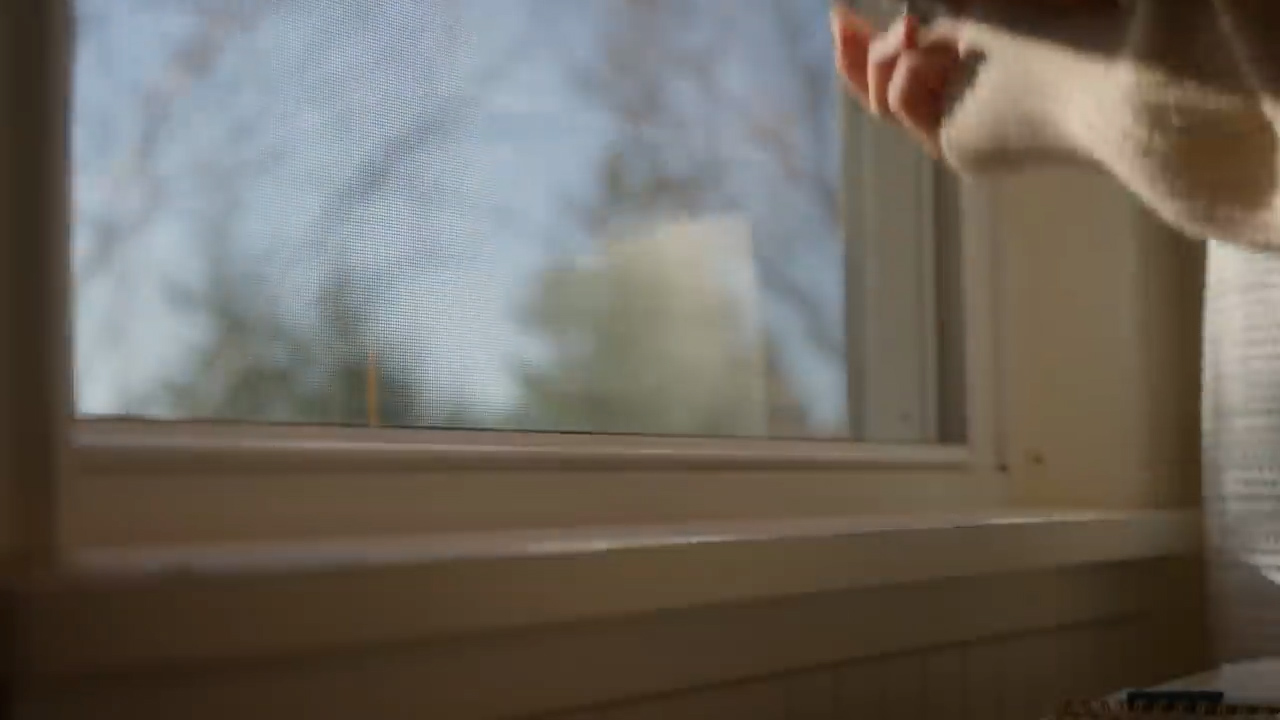 a closeup shot of a hand reaching towards a window. the window has a white frame and a screen, and the sunlight is shining through, casting a warm glow on the scene. the reflection of trees and the sky can be seen in the window. the overall feeling is one of peace and serenity.