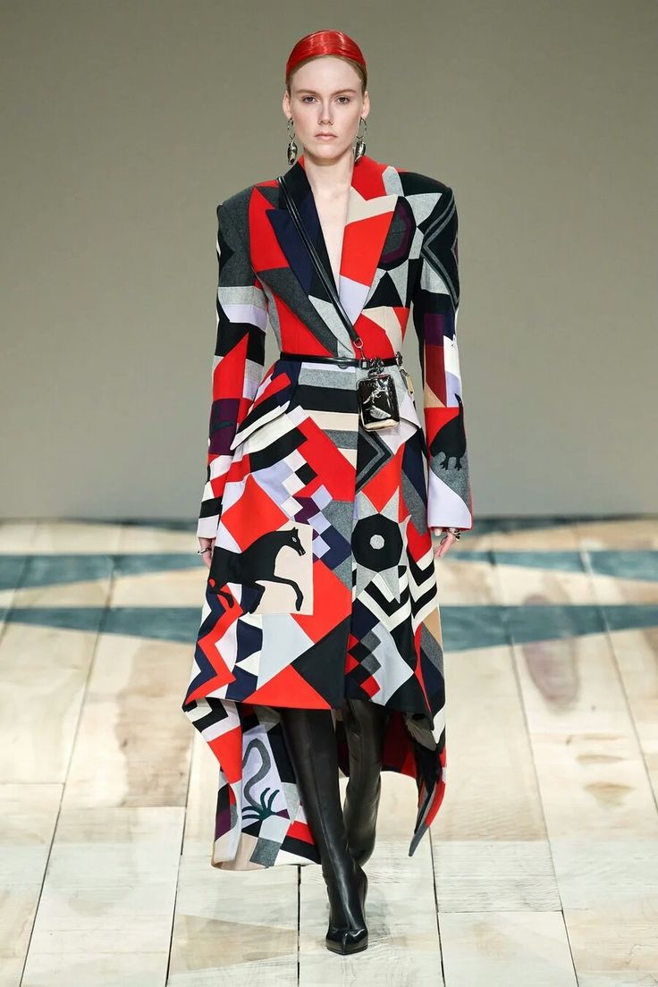 a model walks the runway in a vibrant red and black geometric patterned coat. the coat has a highlow hemline, with a long front and a shorter back. she is wearing black kneehigh boots and a black belt with a small silver bag attached. the model has red hair and wears silver earrings. the background is a patterned floor typical of a fashion runway.  the image captures the energy and excitement of a fashion show, showcasing the latest trends in clothing and accessories.