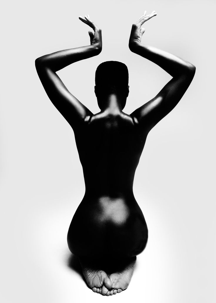 the image is a striking black and white photograph of a nude woman from behind.  her arms are gracefully raised above her head, her hands nearly touching. her body is presented in a contemplative pose, slightly hunched forward, with her feet together on the ground. the lighting is dramatic, creating strong contrasts and emphasizing the curves and form of her body.  the background is plain white, drawing all attention to the subject. the overall aesthetic is minimalistic and artistic, conveying a sense of elegance, serenity, and powerful femininity.