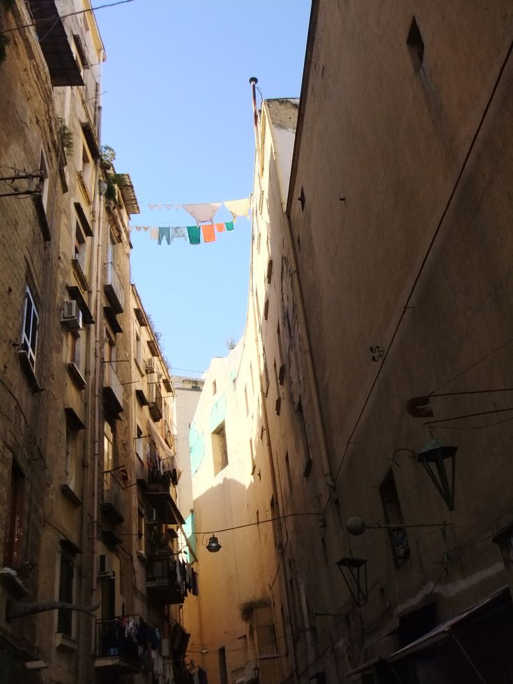the image shows a narrow, sunlit alleyway between two tall, aged buildings.  clothes are hung out to dry on a clothesline stretched between the buildings. the buildings are typical of old italian architecture, showing signs of age and wear.  the scene is quite intimate, showing the close proximity of the buildings and a glimpse into the daily life of the residents. the sunlight casts shadows across the buildings, adding to the atmospheric quality of the photograph.  streetlights are visible along the alleyway.