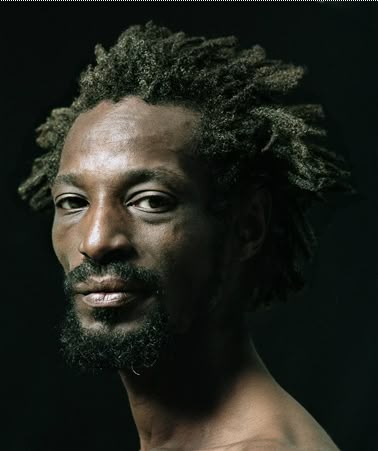 a headshot of a man with dreadlocks and facial hair against a dark background. he is looking at the camera with a serious expression.