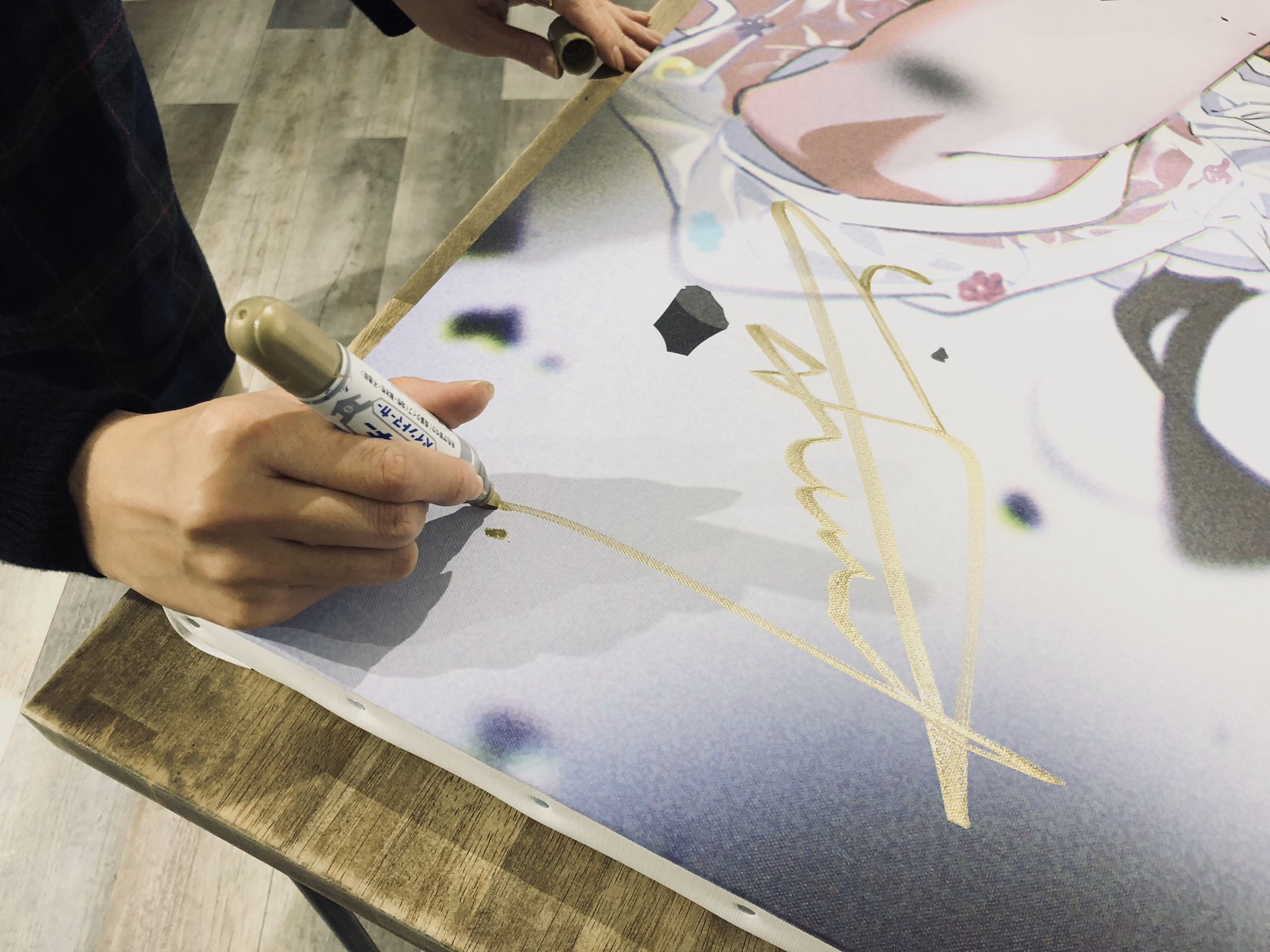 a person is signing their name in gold on a large printed canvas featuring an animestyle illustration. the canvas is resting on a wooden table. the signature is stylized and somewhat difficult to read.