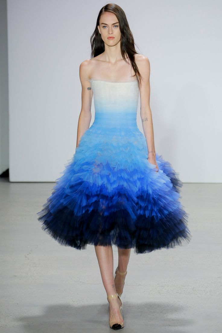 a woman wearing a blue ombre tulle gown walks down a runway. the dress is a light, airy design, with several layers of tulle, creating a soft and romantic silhouette. the model's hair is dark and long, her makeup is natural, and she carries a confident and elegant demeanor. the backdrop is a simple white wall, allowing the dress to be the focus of the image. the overall aesthetic is luxurious and feminine.