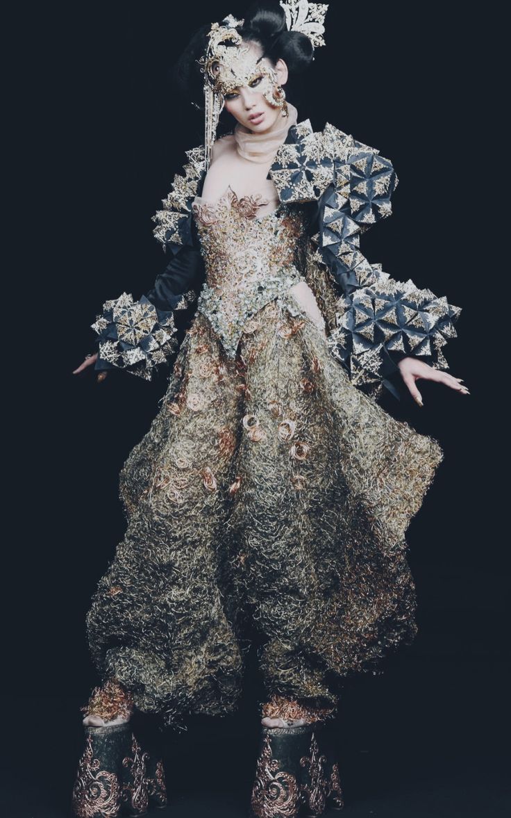 a woman in an elaborate, avantgarde costume poses against a dark background. she wears a heavily embellished dress with intricate detailing, a complex headdress, and sculptural, textured sleeves. the outfit gives off a theatrical and artistic vibe, suggesting a performance art piece or a conceptual fashion statement. the costume's design is unique and extravagant, utilizing texture and embellishment for a dramatic effect. the woman is posed in a fullbody shot, emphasizing the overall impact of the ensemble.