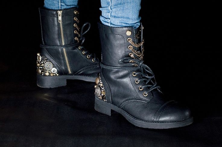 a closeup shot showcases a pair of black leather combat boots adorned with intricate gold and metallic embellishments, including buttons and chains. the boots are laced up and feature a side zipper.  they are being worn by a person whose legs are visible, clad in blue jeans. the background is a stark black, emphasizing the detail of the boots. the overall style is edgy and fashionable.