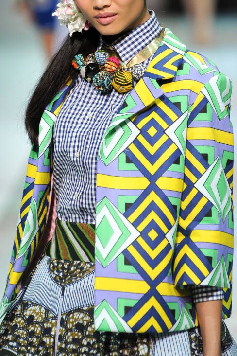 loseup shot of a fashion model showcasing a vibrant outfit. the model is wearing a colorful geometricprinted jacket over a gingham shirt. the jacket features a bold pattern in yellow, green, blue, and purple.  a large statement necklace with colorful spherical beads adds to the eclectic look. the model's hair is pulled back, and a small flower is visible near her ear. the overall aesthetic is bright, summery, and bold.  the background is blurry, suggesting a runway or fashion show setting. the skirt is also visible, with a coordinating print.