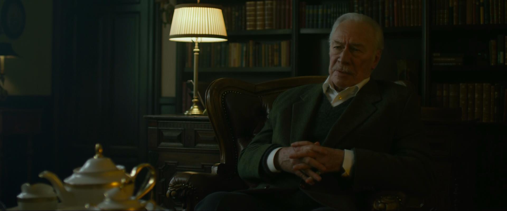a man in a dark suit is sitting in a dark room, looking thoughtful. there's a teapot and a cup of tea in the foreground and a lamp in the background. the man is sitting in a chair next to a bookshelf filled with books. the room is dark and the only light source is the lamp.