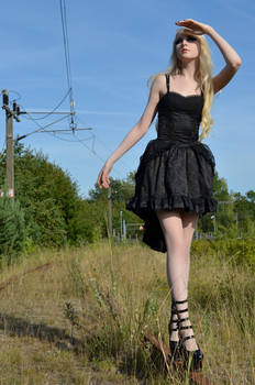 a young blonde woman in a black gothic lolitastyle dress poses outdoors near a railway track.  she is shielding her eyes from the sun and is wearing highheeled boots.  the setting is rural and sunny.