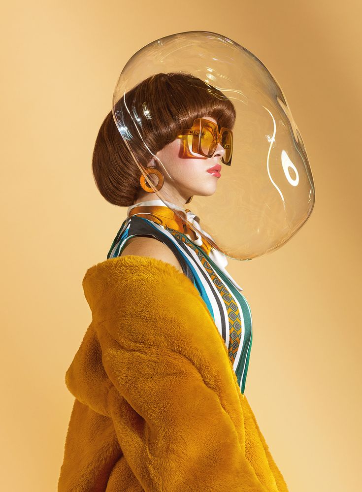a woman in a yellow fur coat and oversized sunglasses poses for a studio portrait with a large, clear plastic bubble around her head. she has short brown hair, and the background is a pale yellow. the woman is wearing green and orange patterned clothing inside the fur coat. her outfit and the bubble add a whimsical and futuristic element to the photo.