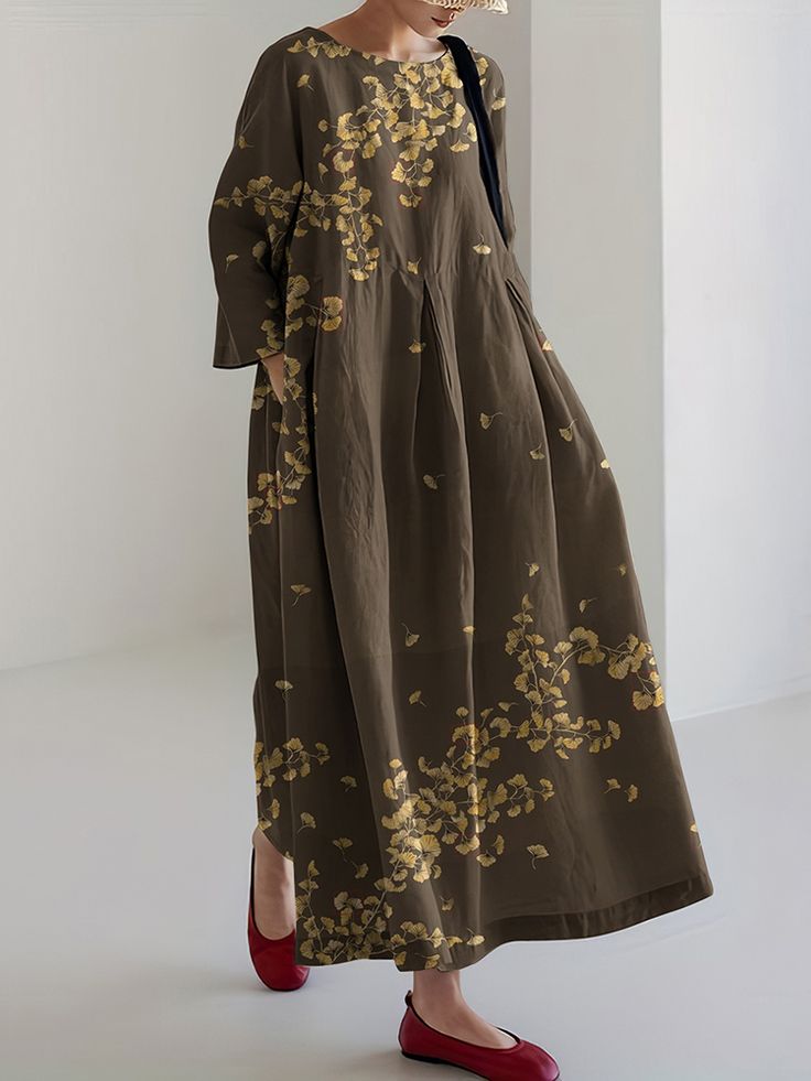 a woman wearing a long brown floral dress and red shoes. the dress is a maxi dress with long sleeves and a round neckline. the floral design is of ginkgo leaves. she is wearing a straw hat. the image is taken from a side view. the background is a plain white wall. the woman looks elegant and casual at the same time.