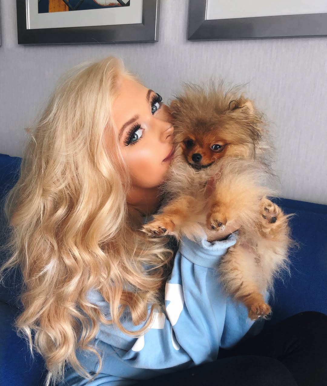 a blonde woman is holding and kissing a pomeranian dog. she is wearing a light blue sweatshirt and sitting on a blue couch. there are two framed pieces of art hanging on the wall behind her. the woman has long blonde hair and is wearing makeup with prominent eyelashes.