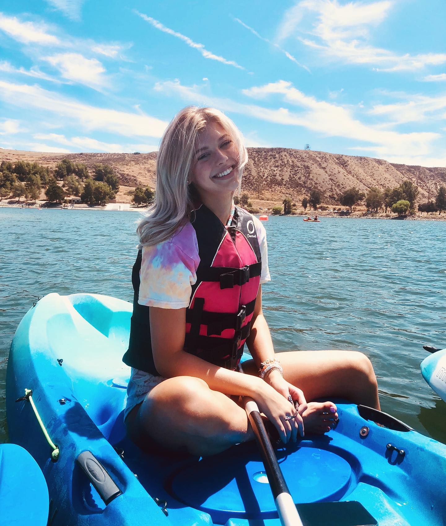 a woman sits in a bright blue kayak on a lake wearing a life jacket and tiedye shirt. she smiles at the camera while holding a paddle. in the background are rolling hills, blue sky and white clouds.