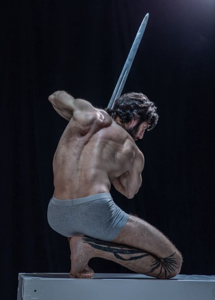 a barechested, muscular man kneels on a white surface, holding a large sword against his back. he wears gray underwear and has a tattoo on his leg. the background is black.