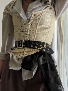 a woman is shown from the chest down wearing a white corset top with a laceup front over a white billowy shirt. she is also wearing brown pants and a thick black belt with metal grommets and a gold chain. she has a black sheer fabric tied around her waist and draped down.