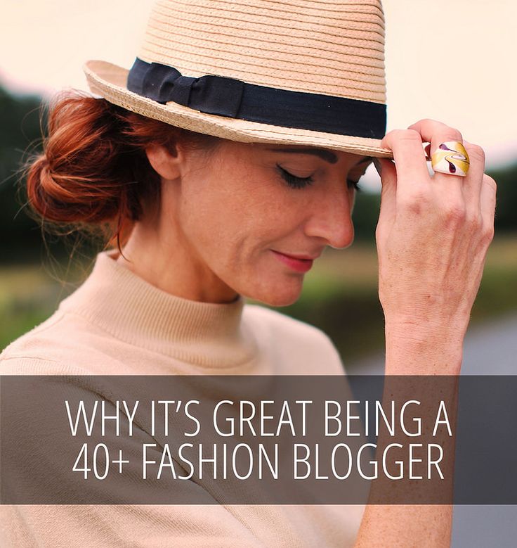 a woman with red hair wearing a beige turtleneck and a straw hat with a black ribbon is posing for a photo. she has a large, colorful ring on her finger and is looking down with a slight smile. text overlaid on the image reads "why it's great being a 40+ fashion blogger". the background is blurry and appears to be outdoors.