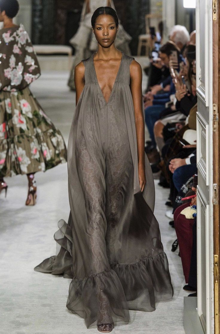 a fashion model walks down the runway in a long grey gown with ruffles. the gown is made of a sheer fabric and has a low neckline. the model is wearing her hair pulled back and has a natural makeup look.  the audience can be seen in the background, watching the show.