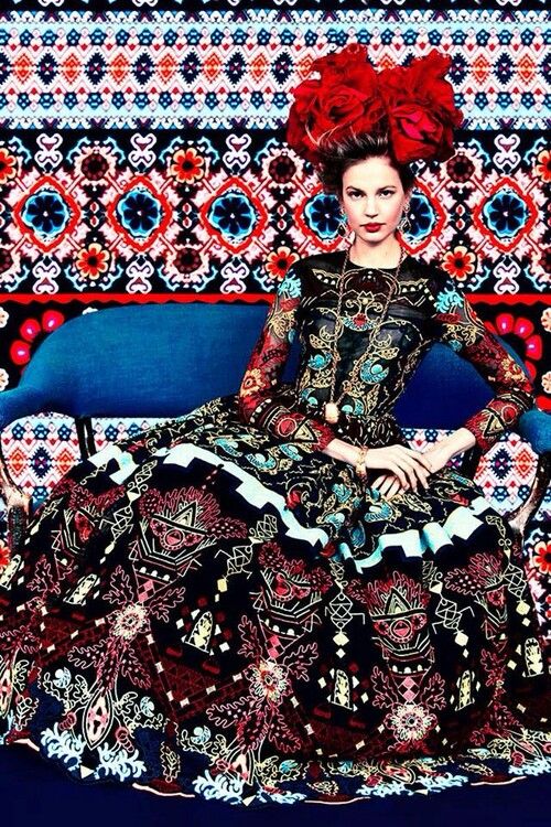 a woman wearing a long black dress with a red headband is sitting on a couch. the dress has a lot of different patterns on it. the couch is blue. the woman is looking to the left of the camera.
