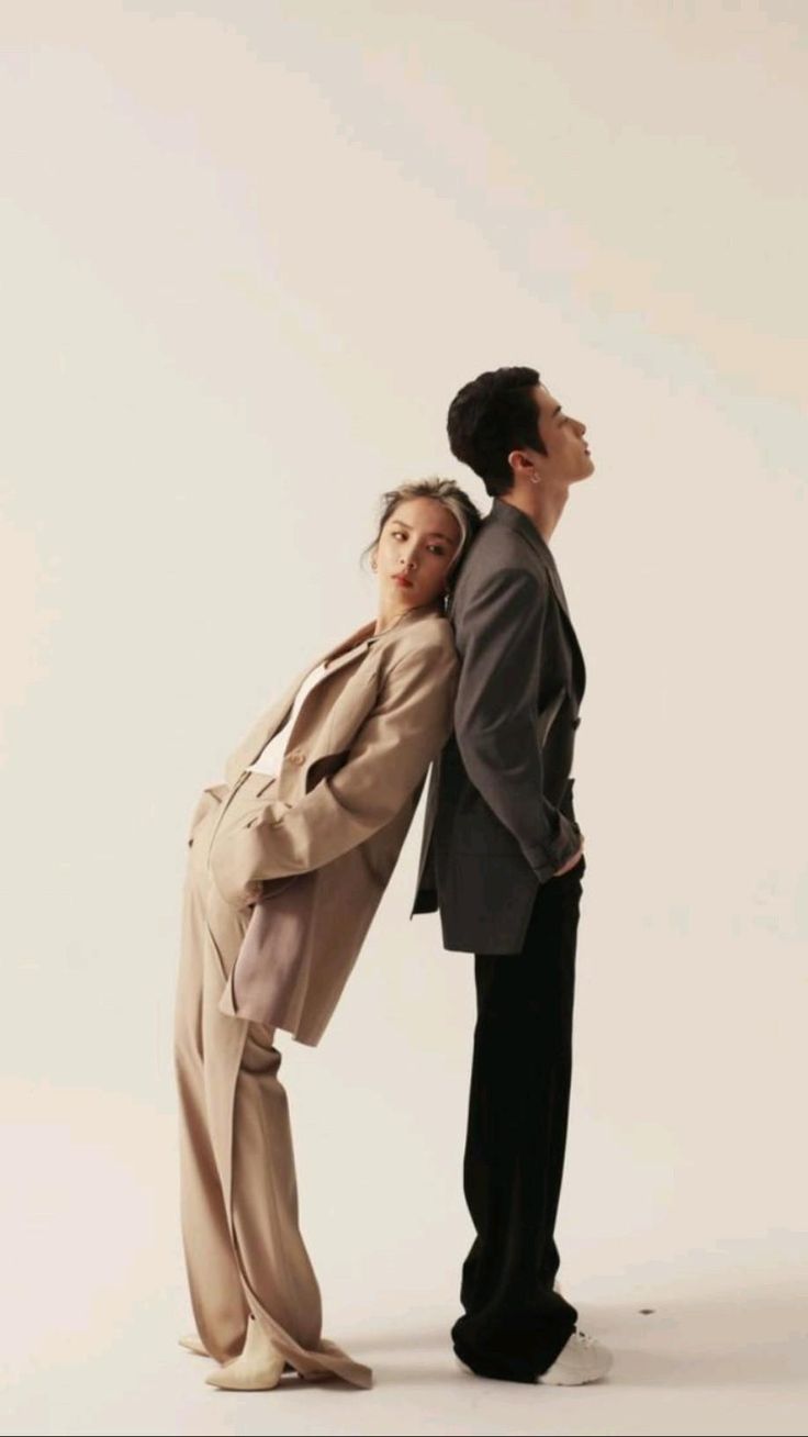 a man and a woman are posing in a studio. the woman is wearing a beige blazer, white top, and beige trousers with white shoes. the man is wearing a black blazer, black trousers, and white shoes. they are standing side by side with their arms around each other.