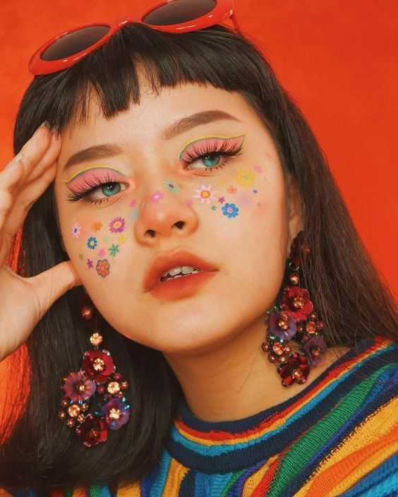 a closeup portrait of a young woman with vibrant and playful makeup. her look features colorful eyeshadow, strategically placed flower stickers resembling freckles, and striking flowershaped earrings. she's wearing red sunglasses and a brightly colored rainbow striped sweater,  creating a retroinspired aesthetic against a solid red backdrop.  her expression is serious and slightly pensive. the overall style is bold, colorful, and reminiscent of 70s or 90s fashion trends.