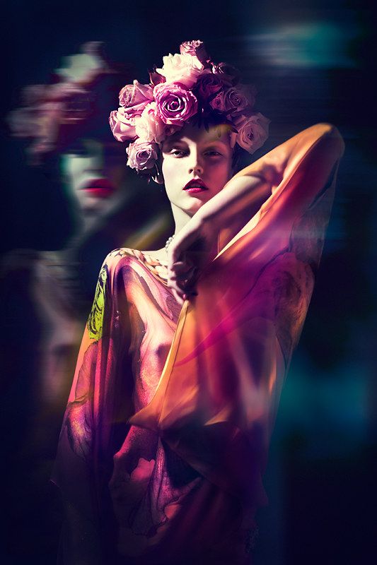 a young woman with a floral crown on her head, wearing a colorful dress, poses dramatically in a dark studio setting. she is illuminated by a soft light, and her expression is intense and mysterious. the image has a glamorous and sensual feel.