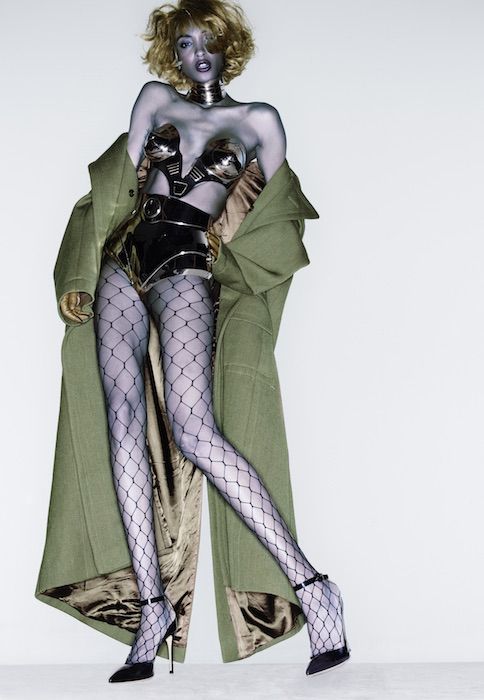 a fashion model is posing in a studio against a white background. she is wearing a green coat, fishnet stockings, a silver bra, a silver belt, and black heels. the model's left leg is slightly bent and her right leg is straight. she is looking at the camera.