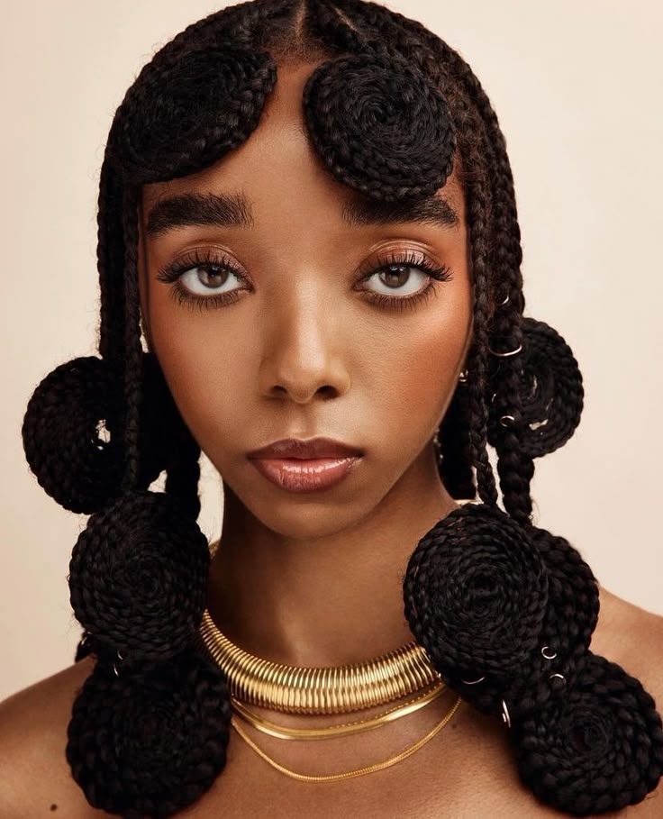 the image is a portrait of a woman with an elaborate braided hairstyle. the braids are styled into circular shapes around her forehead and along the sides of her face. she has dark skin, brown eyes, and glossy lips. she is wearing two gold necklaces, one thick and one thin. the background is a neutral color.