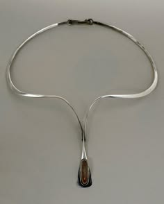 a silver necklace with an oval pendant that contains a gemstone.  the necklace has a unique shape, with two thin silver bars that extend down from the choker and meet in a single bar that ends at the pendant.  the pendant is oval shaped with a round, amber colored gemstone.  the pendant is slightly wider at the top than at the bottom. the necklace appears to be handmade and has a rustic look to it. the necklace appears to be silver but it is impossible to tell the exact metal without more information. the pendant is made of silver, and the gemstone is oval shaped and sits in a silver bezel setting. the necklace is elegant and simple, yet striking. the necklace would be a great gift for someone who enjoys unique and modern jewelry.