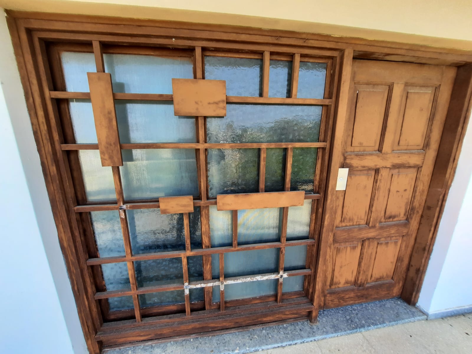 an eyelevel, slightly skewed shot of a wooden window and door combination. the window is covered with a grid of wooden bars and panels, while the door is paneled. the wall to the side of the window and door is white. the overall image has a slightly rustic and worn appearance.