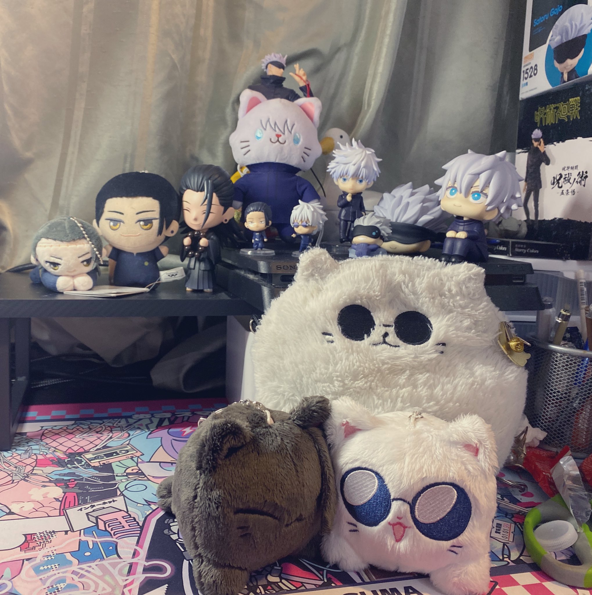 a collection of anime and mangathemed plush toys and figurines are displayed on a table. the plush toys include a white cat with black eyes, a white cat with blue sunglasses, and a brown bearlike creature. the figurines include a variety of characters, mostly in black and white outfits. the background is a blurred image of a wall and a bookcase. the overall scene is bright and playful.