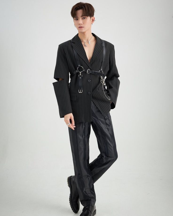 a full shot of a man posing in a stylish black pinstripe suit with a harness detail. he is also wearing earrings, a ring, and a necklace. the outfit is completed with black boots.